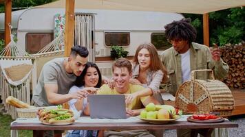 A diverse group of friends relaxes outdoors in a camping setting, sharing laughter while looking at a laptop, have call. The atmosphere is filled with joy and camaraderie on a warm summer day. video