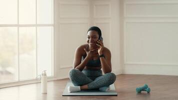 In a bright indoor space, a young African American woman sits cross-legged on a workout mat, smiling while chatting on her phone. She's embracing a healthy lifestyle and fitness routine. video