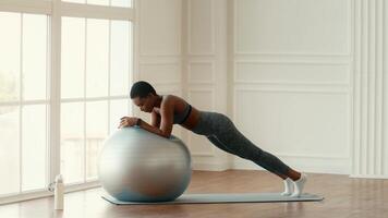 A woman engages in a plank exercise using a stability ball at home. She maintains focus while practicing an athletic routine in a bright and spacious indoor area, promoting a healthy lifestyle. video