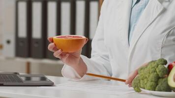 A healthcare professional in a white coat is demonstrating food preparation with fresh ingredients like broccoli and orange while discussing dietary choices with clients in a clinic. video
