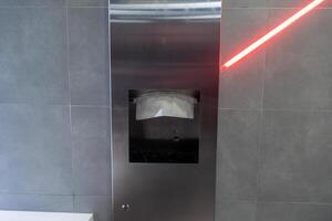 A metallic sanitary disposal unit is mounted on a wall with a red laser indicator, highlighting its functionality in a sleek, modern restroom setting photo