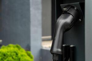 A charging connector hangs from an electric vehicle charging station. Bright green foliage is visible nearby, creating a clean, eco-friendly atmosphere in an urban area photo