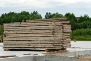 A stack of wooden planks is neatly arranged on a concrete surface at a construction site surrounded by lush greenery under a cloudy sky photo