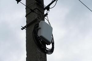 Communication hardware is mounted on a utility pole with wires hanging down. The backdrop features a gray, cloud-filled sky indicative of overcast weather photo