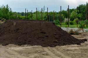 A sizable pile of dark soil sits on a farm landscape surrounded by patches of grass and emerging vegetation under a cloudy sky in a rural setting photo