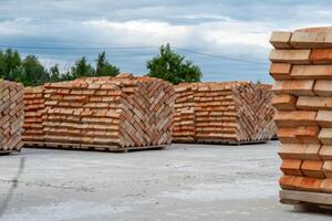 Piles of neatly arranged red bricks sit in a construction yard, ready for use. The sky is overcast, hinting at possible rain. Green trees provide a backdrop photo