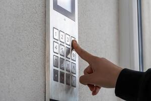 A person uses their finger to press a number on an intercom system located at the entrance of a residential building during the day. The wall is smooth and light-colored photo