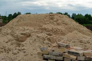 A significant mound of sand rises prominently beside neatly stacked bricks on a construction site. Overcast skies loom above, hinting at potential rain photo