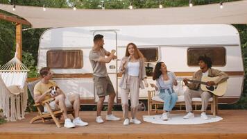A diverse group of friends is having fun during a summer weekend camping trip. They are singing and dancing together by a vintage trailer, surrounded by nature. video