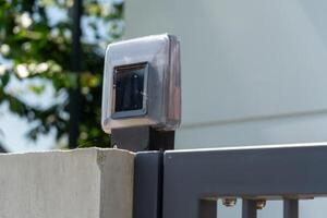 A solar-powered light mounted on a gray metal gate provides illumination and security. It is situated in a sunny outdoor area with greenery in the background photo