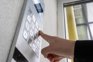 A person is pressing a button on a sleek security entry system attached to a wall in an urban setting while sunlight filters through nearby windows photo
