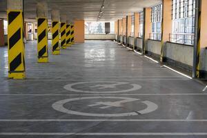 A spacious parking garage features clear directional markings with yellow stripes. Natural light filters in through large windows, creating a bright atmosphere photo