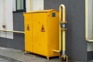 Utility cabinet painted in bright yellow is placed beside a gray wall, featuring warning signs. Pipes run along the structure, indicating safety measures and functionality photo