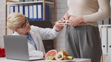 A dietitian takes body measurements of a client while discussing healthy food options. Fresh fruits are displayed on the table, showcasing a balanced approach to nutrition in a clinic. video
