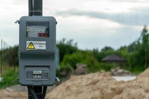 A new electrical meter box is mounted on a pole at a construction site surrounded by sand and greenery under overcast skies photo