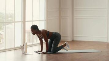 An African American woman engages in online yoga session at home, focusing on her fitness routine. The spacious room features natural light filtering through large windows. video