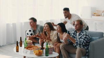 A group of diverse friends relax at home, cheering enthusiastically while watching a match. They share snacks and drinks, embracing a lively, fun atmosphere together. video