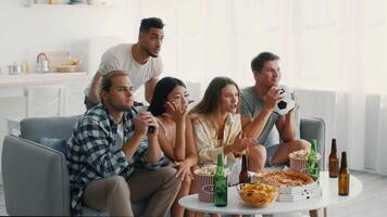 A group of diverse friends are gathered in a cozy living room, enjoying snacks and drinks while watching a sports event on television. The atmosphere is filled with excitement. video