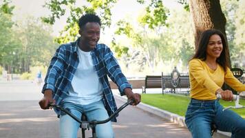 A joyful couple rides bicycles through a picturesque park, laughter filling the air as they enjoy the sunny weather. Friends are seen nearby, creating a vibrant outdoor atmosphere. video