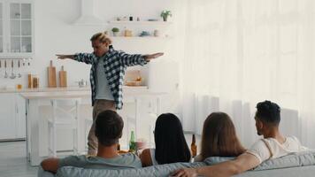 A group of diverse millennials gathers in a bright home, sharing laughter and good times. One person stands and entertains while friends relax on the couch. video