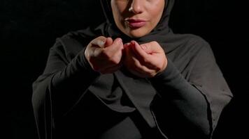 woman wearing black hijab sits quietly her hands held together. pours black sand, setting is dimly lit creating serene atmosphere for introspection video