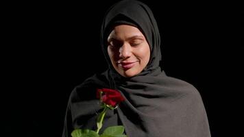 woman wearing black hijab stands quietly closed eyes gently holding red rose and smell it. background is dark emphasizing her peaceful expression and connection flower video