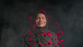 woman in black hijab stands serenely her eyes closed as vibrant red rose petals cascade around her in smoky backdrop creating peaceful atmosphere video