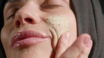 In cozy indoor setting woman wearing hijab gently applies green skincare product her cheek focusing on her self care routine. light creates serene atmosphere video