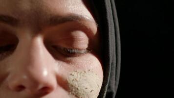 woman in black hijab is engaged in applying artistic facial makeup. focus is on her face showcasing intricate details and textures. setting is illuminated soft daylight video
