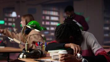 africano americano aluna queda adormecido em livros pilha dentro a campus biblioteca, lidando com esgotamento e lutando para graduado faculdade. jovem aluno com fadiga tentativas para estude às noite. Câmera uma. video