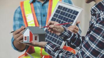 Two men are holding a model of a house with a solar panel video