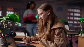 Faculdade aluna trabalhando em computador portátil debaixo verde luminária dentro uma quieto contexto, usando universidade biblioteca dentro a campus para preparar para importante escola projeto. jovem menina reúne dados. Câmera uma. video