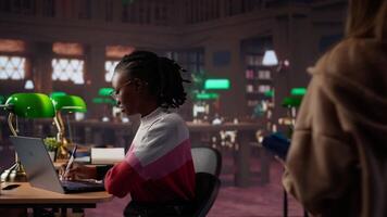 University student making exam preparations with a friend at the campus library, doing research to complete handwritten class notes. Girls sharing information from the database. Camera A. video