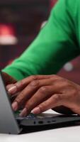 Vertical African american freelancer at home office sitting at desk and typing on laptop keyboard, writing down ideas to solve his daily assignments. Remote worker answering inquiries. Camera A. video