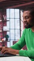 Vertical African american remote worker solving his daily freelancing tasks on laptop, working in relaxation from the comfort of his modern loft. Tech savvy black man internet user, multitasking. Camera B. video