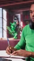 vertical sonriente negro hombre tomando notas mientras acecho un tutorial presentación, haciendo en línea yo aprendizaje y escritura detalles. joven adulto persona de libre dedicación escribe en su libro de texto bloc. cámara b. video