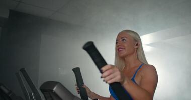 woman is focused on her workout as she exercises on an elliptical trainer in a well equipped gym. She wears headphones that keep her motivated while the ambient light creates a striking atmosphere video
