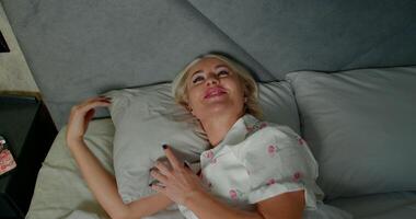 In a cozy bedroom setting late at night a girl rests comfortably in bed with headphones on smiling as she enjoys a phone call. Her relaxed demeanor suggests a friendly conversation video