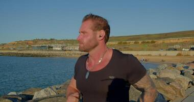 athletic man with a beard jogs confidently along a picturesque seashore while listening to music through his headphones enjoying the sunny weather and ocean breeze video