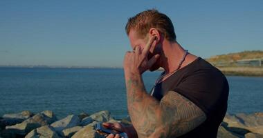 Athletic man with a beard stands on the seashore engaging in a phone call with headphones. The sun shines brightly and the waves gently crash against the rocks nearby video