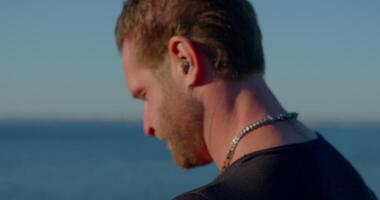 On the peaceful seashore a fit man with a beard is engaged in a phone call using headphones enjoying the scenic view of the ocean and clear sky video