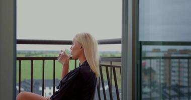 On a balcony a girl with long blonde hair sits thoughtfully while conversing on her phone with headphones. She sips water and enjoys the tranquil view of the green fields video