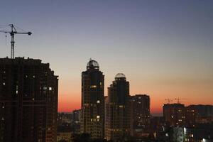 Urban skyline at dusk featuring modern high-rise buildings with illuminated windows, construction crane, and a vibrant sunset sky creating a stunning cityscape atmosphere photo