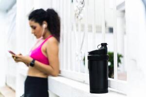 selective focus of a black drink bottle, at the one out of focus woman in sportswear wearing headphones and a phone. photo