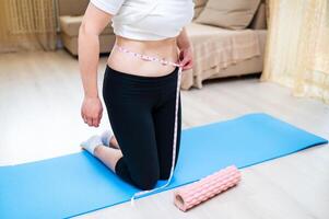 Woman with imperfect waist measuring it with tape while standing on yoga mat, body awareness photo