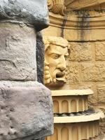 Classical stone sculpture in a historic fountain. The sculpted face, with intricate details, is set against a weathered yellow wall, partially hidden by rough stone blocks. photo