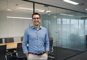 A man in a blue shirt and glasses standing in an office photo