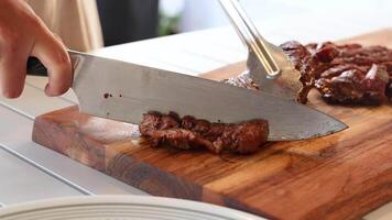 Chef slices cooked beef steak on a wooden board using a large knife and fork. Bright kitchen setting video