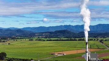 A tall smokestack emits white smoke into a clear sky over expansive green fields, with distant mountains under bright daylight video