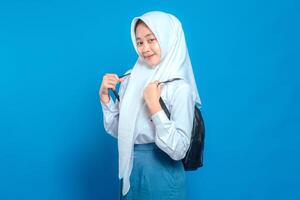 Cheerful Asian teenage girl wearing school uniform and hijab, smiling confidently while holding backpack straps. Positive and friendly facial expression on blue background. photo